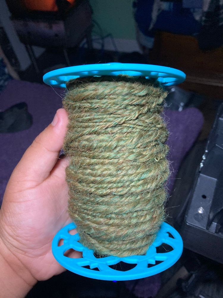 A blue e-spinner bobbin with a three ply green yarn on it.