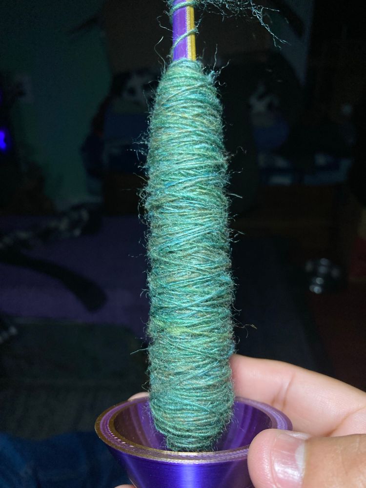 A hand holding a spindle with yarn on it. The yarn is light green. It is a blend of Shetland and Silk