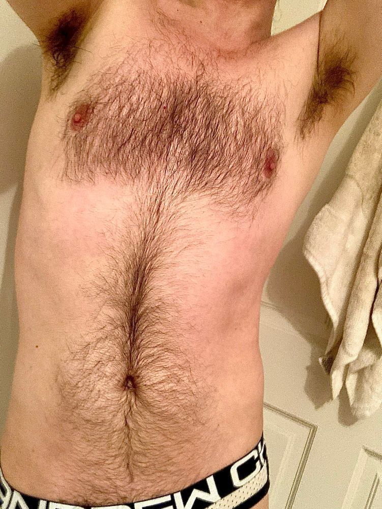 standing in only a jockstrap showing off the fur and pits in bright bathroom overhead lighting 