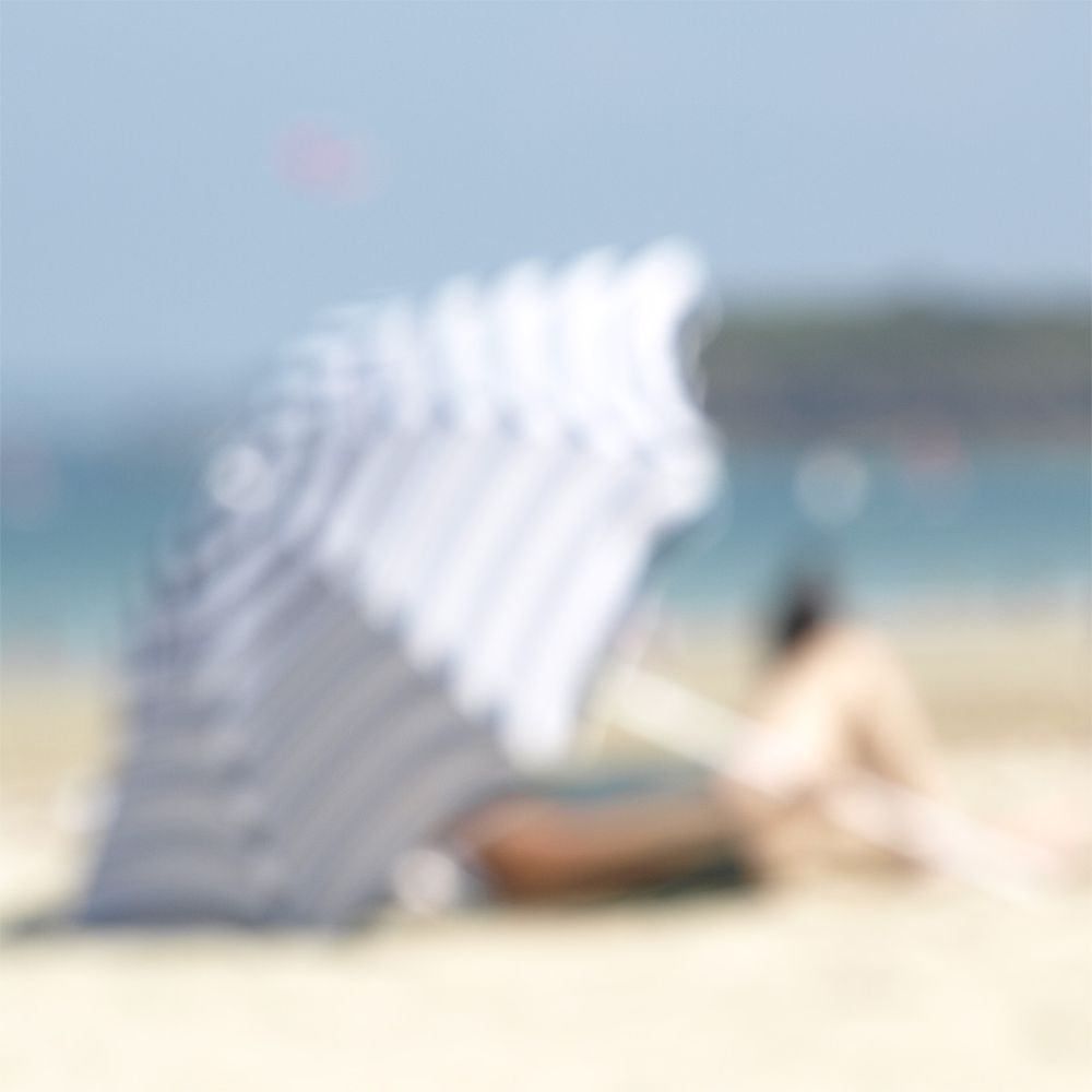 Unscharfe Aufnahme eines gestreiften Sonnenschirms und einer liegenden Person am Strand mit Meer im Hintergrund.