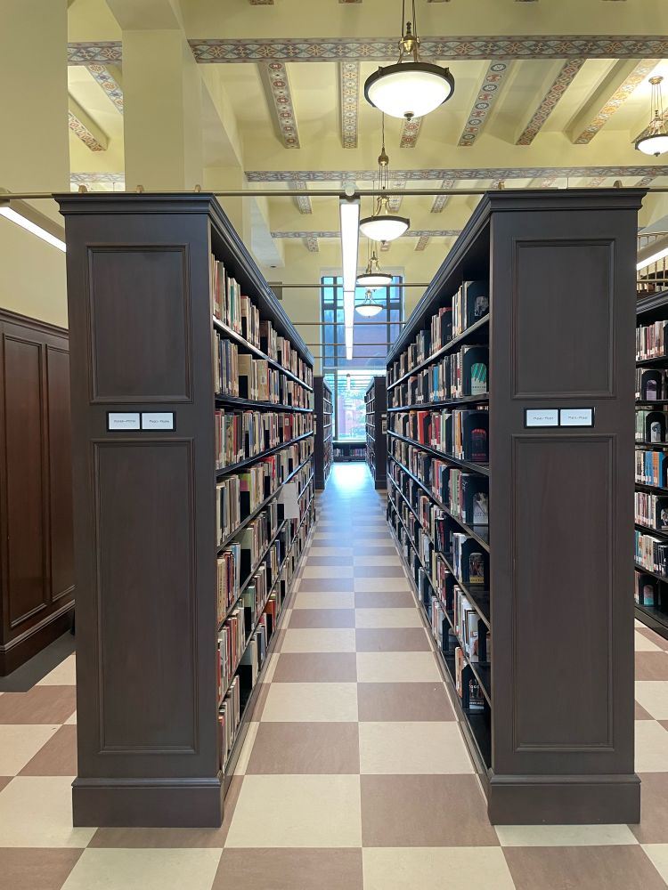 A photo of the Enoch Pratt Free Library in Baltimore. Two long bookshelves stretch away from the camera on a tiled floor. 
