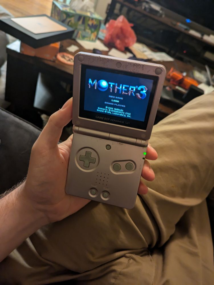 My big hand holding a Game Boy Advanced SP with the Mother 3 title screen displayed. The Coffee Table in the background is cluttered, but not to the extent where it's concerning.