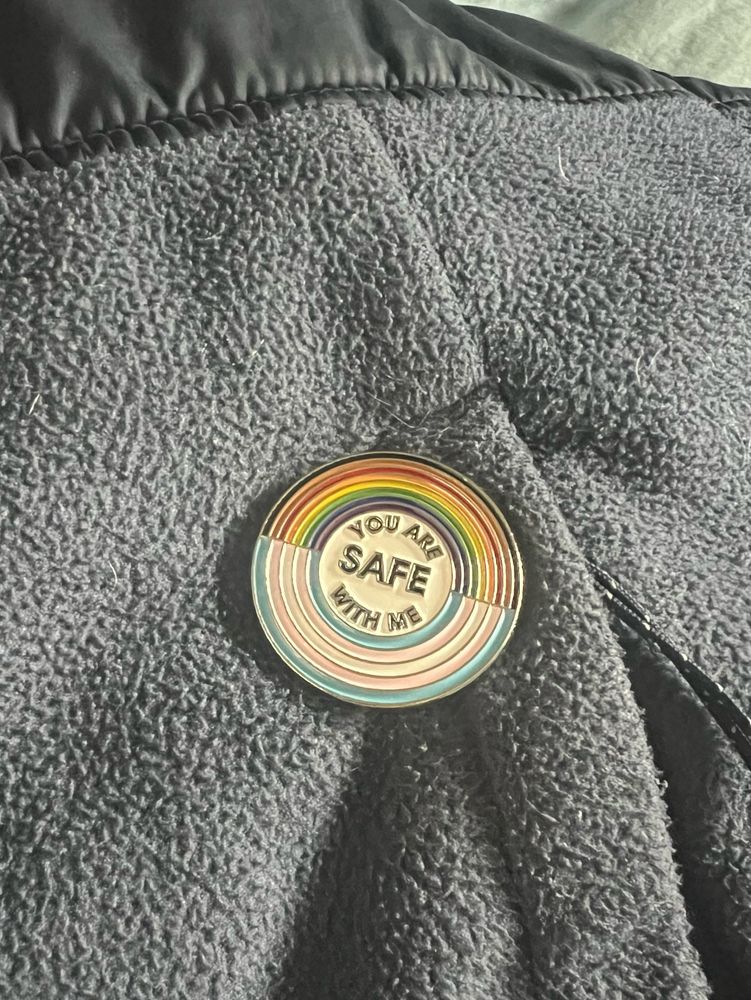 A picture of a round pin on a blue jacket. The top half is a pride rainbow and the bottom is a trans flag representation. In the middle it says "YOU ARE SAFE WITH ME"