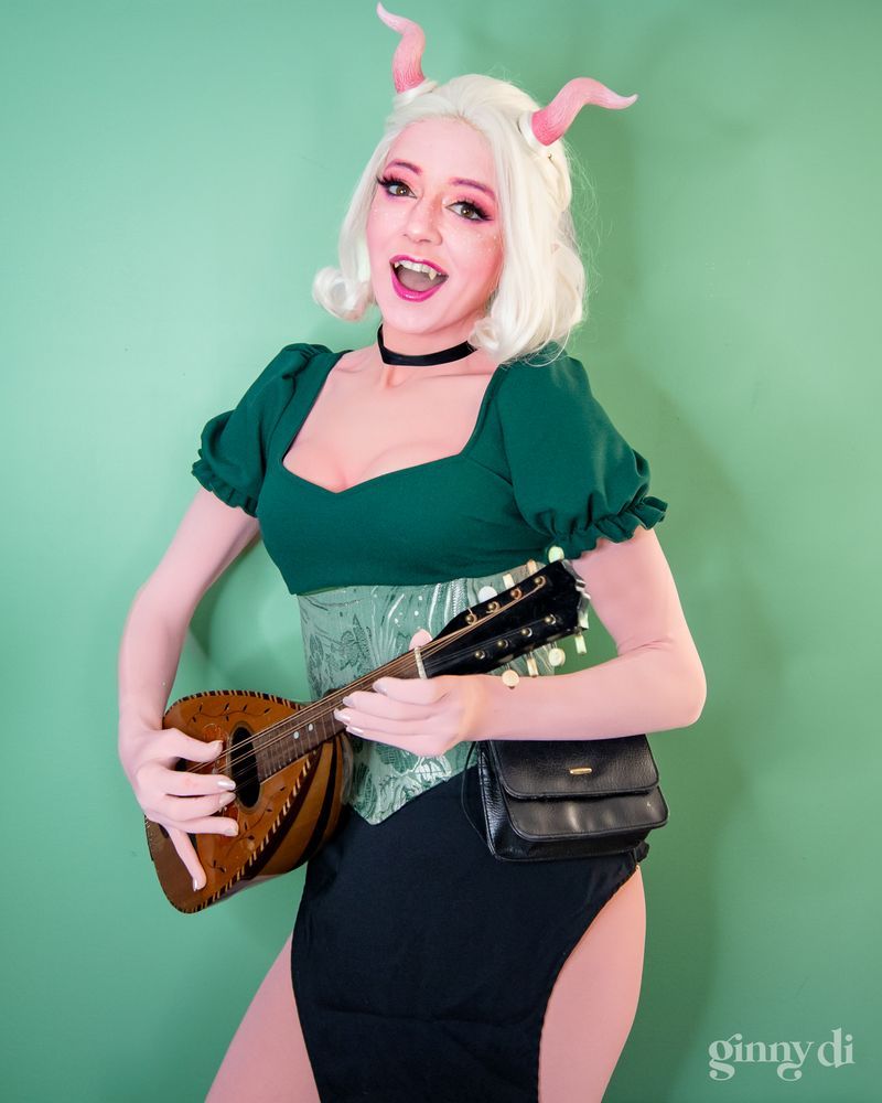 A pink-skinned woman with white hair and short, curled pink horns sings and plays a stringed instrument. She wears a dark green puff sleeve shirt, a green corset, and a black skirt.