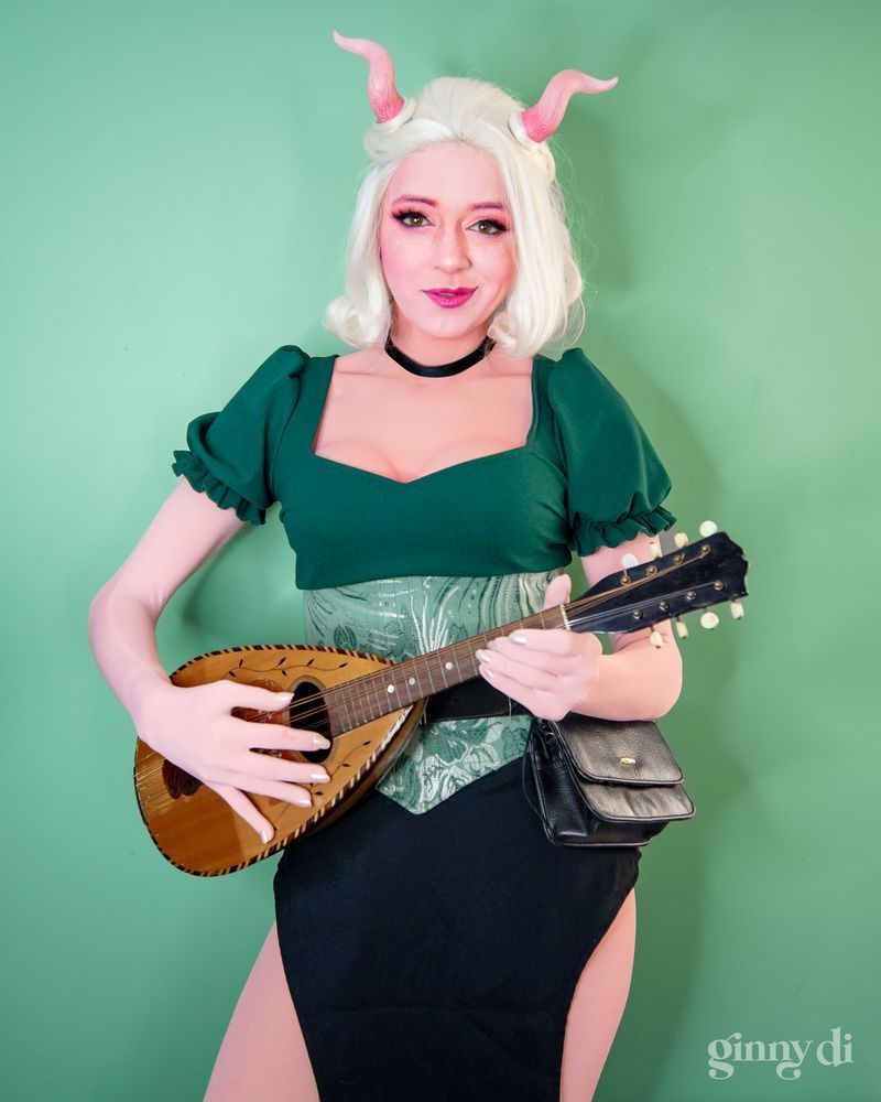 A pink-skinned woman with white hair and short, curled pink horns looks at the camera, smiling and holding a stringed instrument. She wears a dark green puff sleeve shirt, a green corset, and a black skirt.