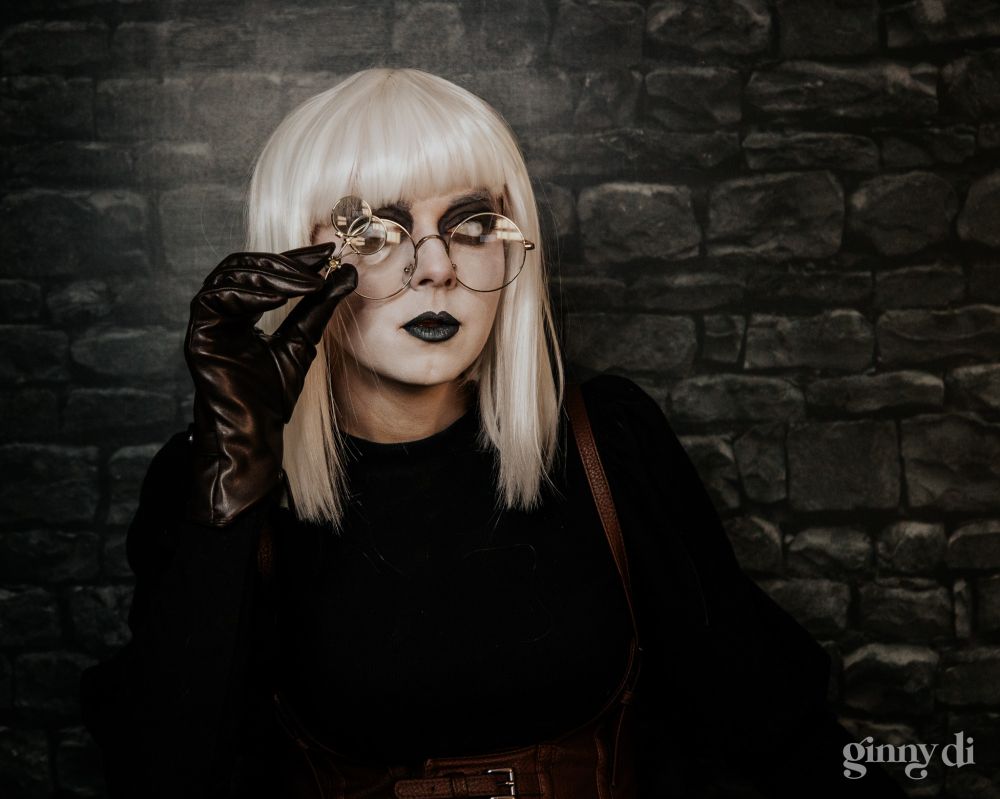 A woman with a blunt white bob, white skin, all-white eyes surrounded by smudged black makeup, and black lipstick. She has a neutral expression. She wears large, round glasses with an attached jeweler's loop, and black leather gloves.