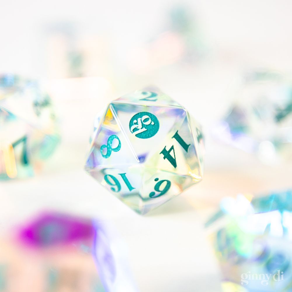 A full set of prismatic glass dice, refracting light in blue, green, yellow, and pink. The other dice are out of focus, but in the center is a d20 in focus. Facing the camera is the 20-face, which has a "gd" logo inked in teal.