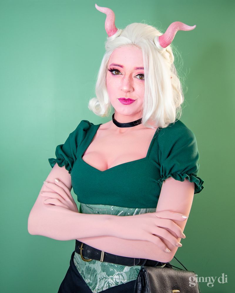 A pink-skinned woman with white hair and short, curled pink horns looks at camera, arms folded. She has white freckles and wears a dark green puff-sleeve shirt and a green waist cincher corset.