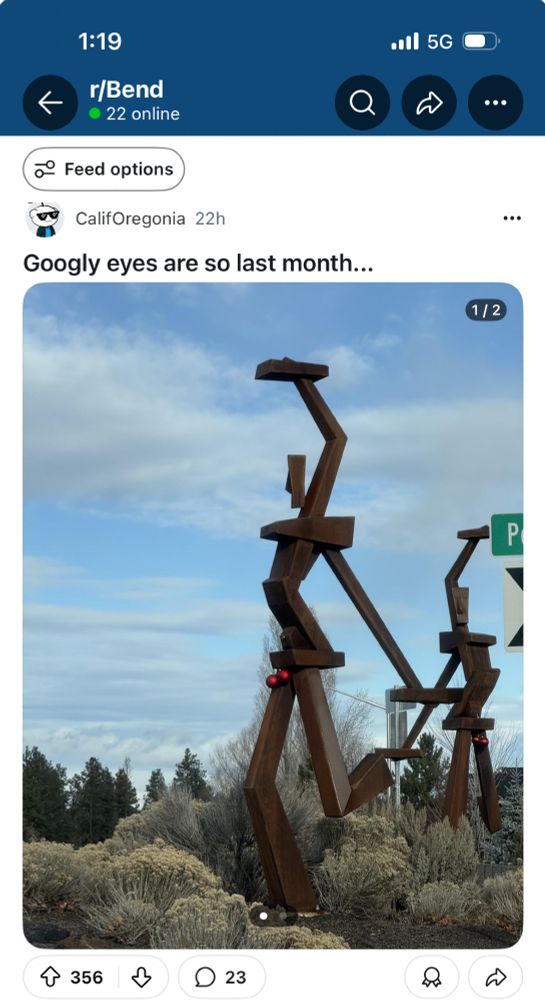 Large abstract sculpture with a faint resemblance to a man has two red Christmas ornaments hanging from the crotch