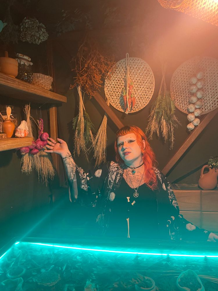 Eu posando numa cabine cheia de ervas e flores secas com uma mão prestes a pegar um potinho. Há uma
mesa bem iluminada com luz verde cheia de objetos como cristais e mais flores secas bem em frente. 