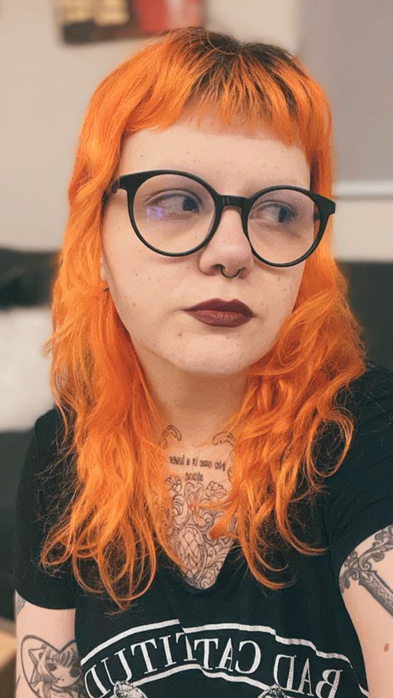 Meowry é branca, tem cabelos longos de cor laranja, usa óculos e batom vermelho escuro. Está olhando para o lado.