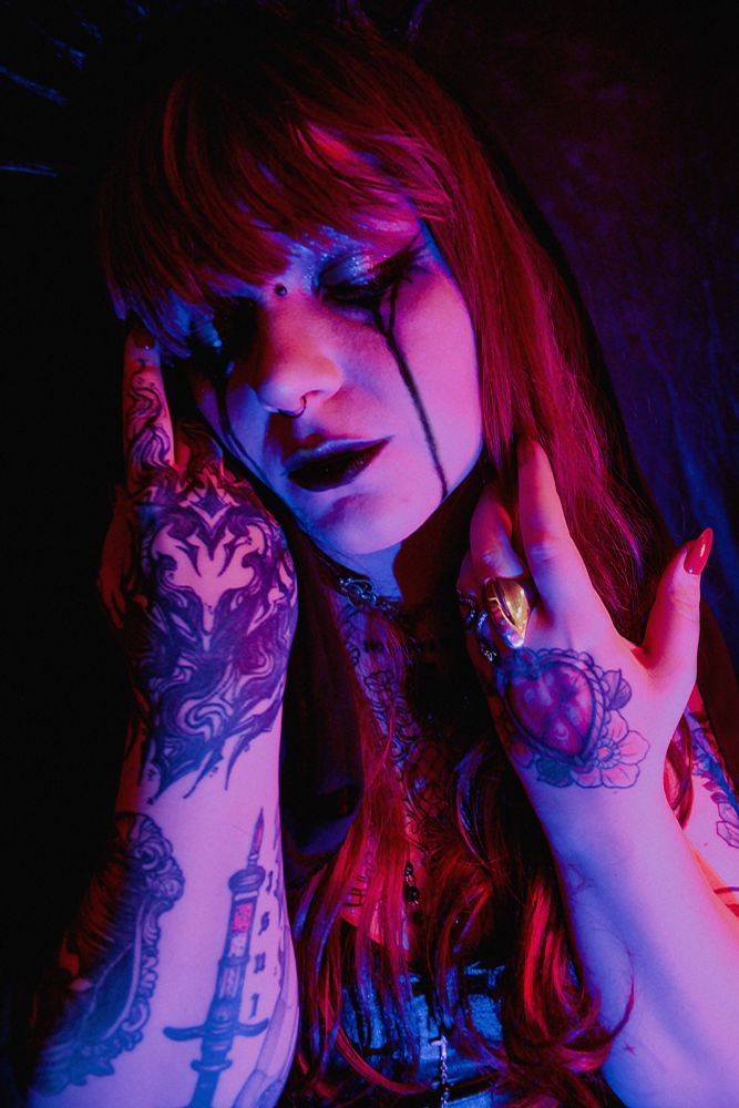 Meowry posando de olhos fechados, usa peruca longa e véu preto sobre a cabeça. A iluminação é roxa. Usa maquiagem preta pesada escorrendo pelo rosto, as mãos estão próximas do rosto, com suas tatuagens visíveis. Sua expressão é sublime.