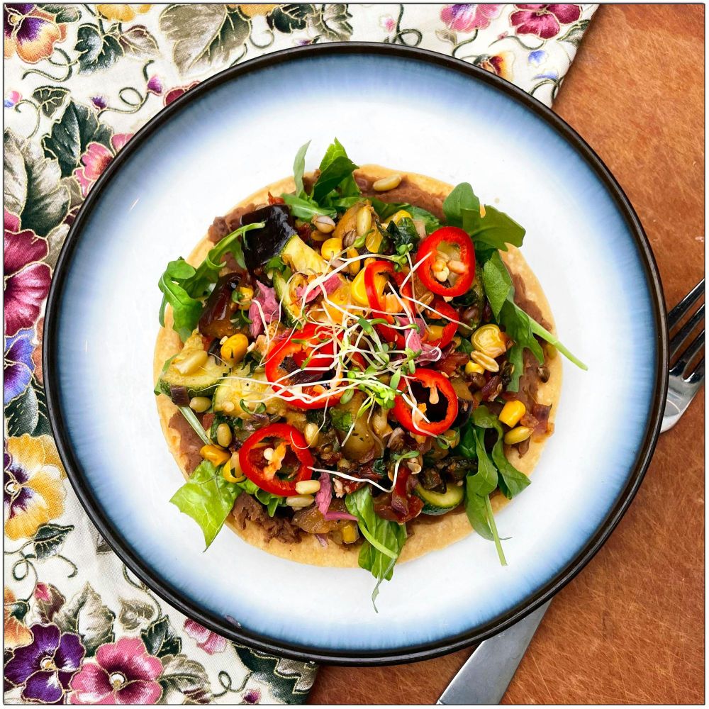 Plate with a round tortilla with "refried" pinto beans, kamut and purple barley, eggplant, zucchini, corn, poblano, and tomatoes lettuce, pickled garlic, microgreens, and sliced hot pepper.