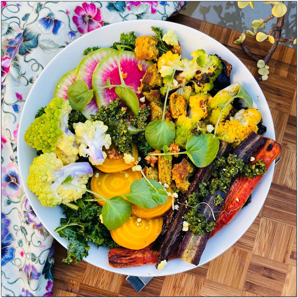 White salad bowl with green romanesco, watermelon radish, golden beets, multi-colored carrots with a carrot pesto topping on a bed of roasted purple cabbage and kale.