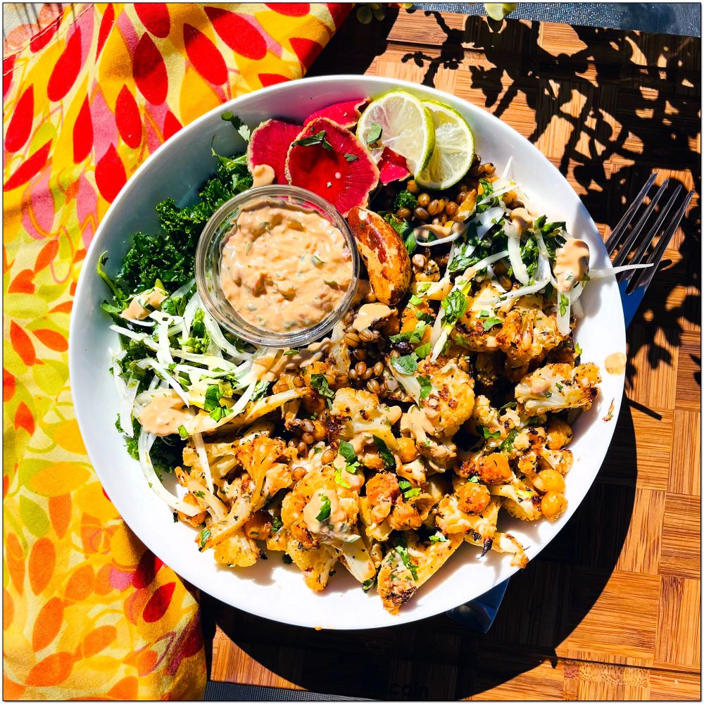 A white salad bowl filled with chickpeas, a brazil nut, purple barley, cauliflower, onion, watermelon radish on a bed of kale.