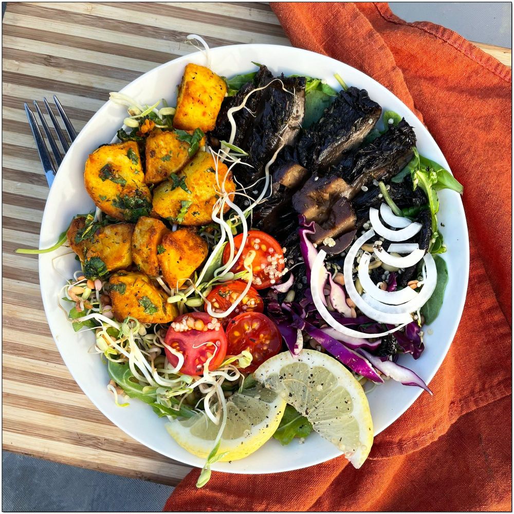 White bowl with yellow potatoes, sliced mushrooms, white onions, tomatoes, purple cabbage, bean sprouts, and slices of lemon on a bed of greens.
