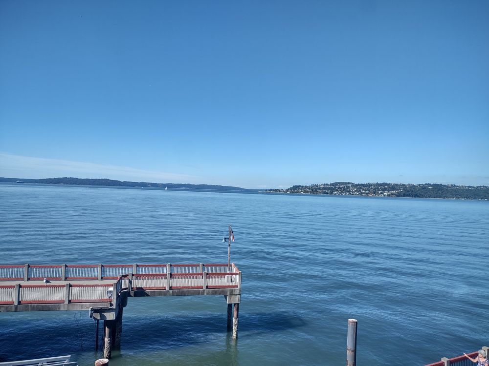 A photo of Commencement Bay. The expanse of water has distant views of Maury and Vashon Islands. A concrete pedestrian pier is visible on the left.