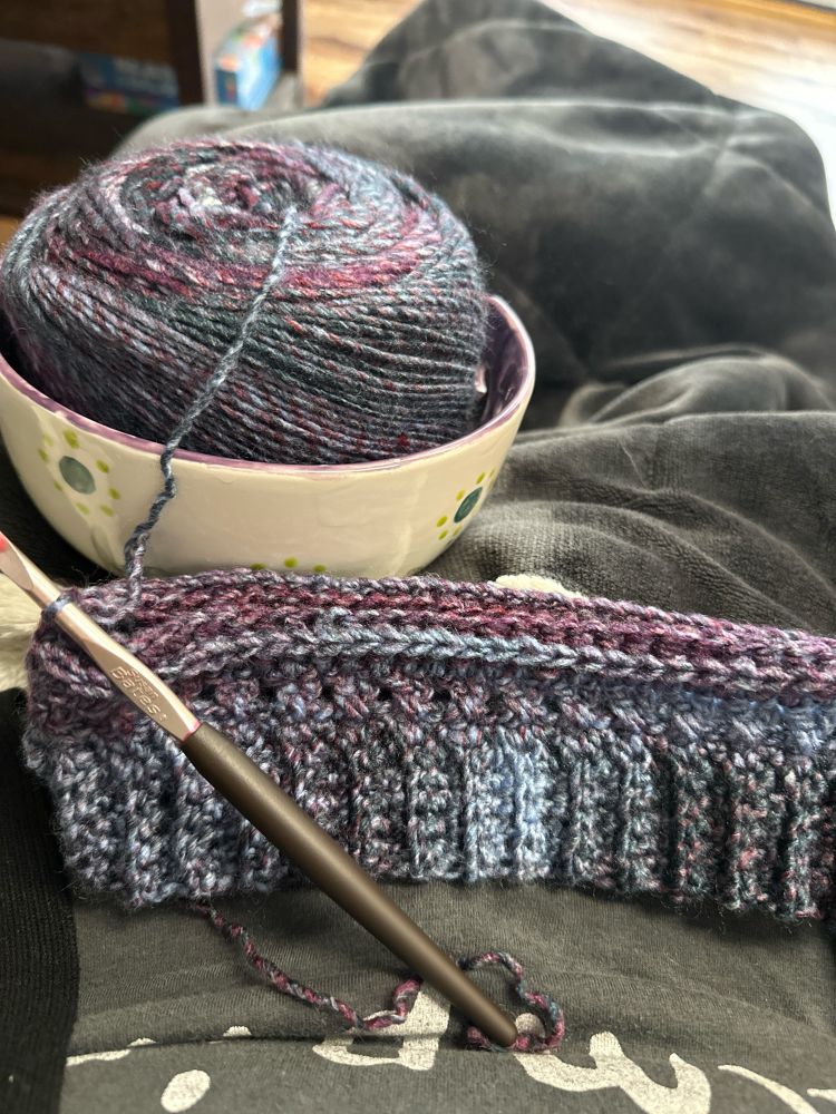 A blue and purple crocheted hat partially finished with a crochet hook and ball of yarn in a yarn bowl next to it.
