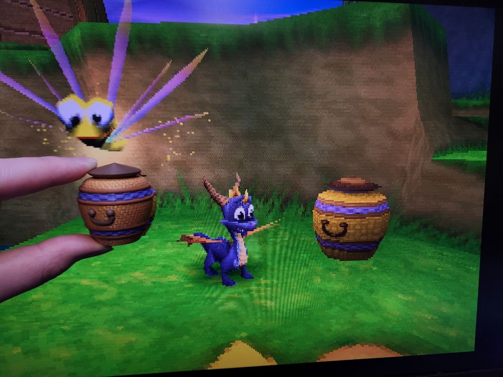 A white hand holds a 3D-printed golden basket with dark brown and purple accents in front of a screen displaying Spyro 3 on the Playstation 1. The basket is held, over top of an in-game one, so that it looks like the hand is grabbing something right out of the game. Spyro the purple dragon is looking away, while his companion Sparx - the yellow dragonfly - is looking at the viewer, seemingly aware of what is happening.