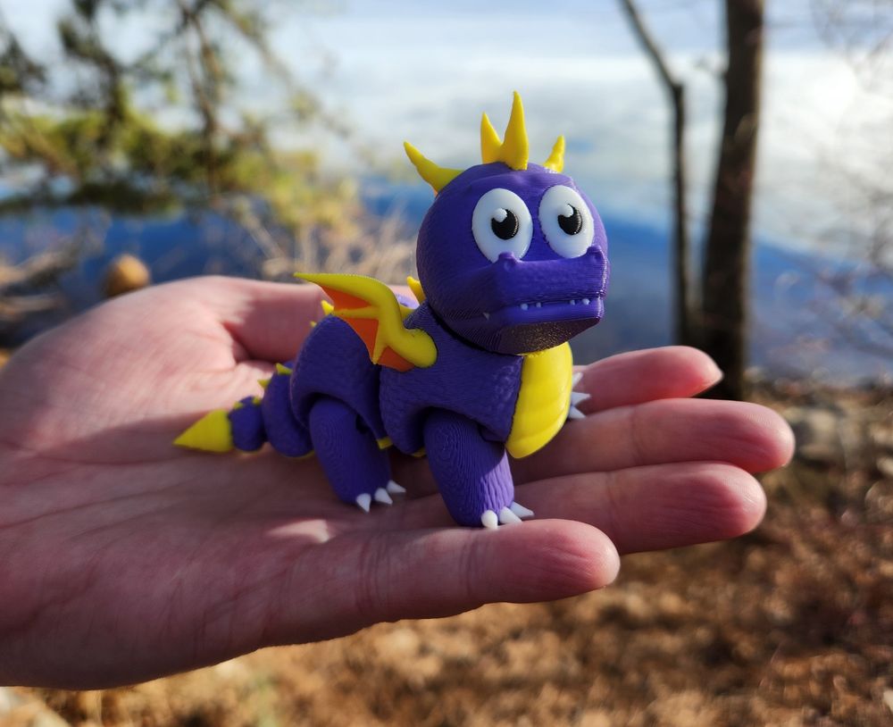 A white hand holds a 3D-printed toy of a baby version of Spyro the Dragon. Spyro has a purple body with yellow features (belly, horns, spikes, and wings) and orange webbing for the wings. He has moving parts, and an oversized head, with a funny look to his face. In the background are some trees and the edge of a partially-frozen body of water.