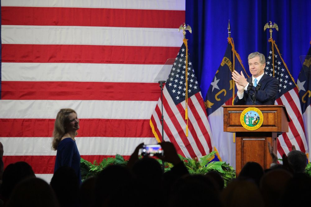 North Carolina Gov. Roy Cooper, standing behind a podium flanked by American flags, leads a round of applause for First Lady Kristin Cooper (standing to the left in the audience) during his farewell address at Nash Community College in Rocky Mount.
