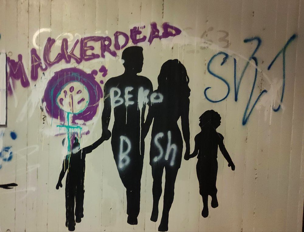 Ein Graffiti mit dem Schriftzug ,Macker Dead' über einer Familie mit 2 Kindern in schwarzer Silhouette. Das Graffiti selbst ist Lila, Weiß mit einem Schnörkel und mit Türkis und Pastell-Orange inklusive einem Feminismus*-Zeichen gefüllt und markiert. Am linken Rand eine weiße Leiste.