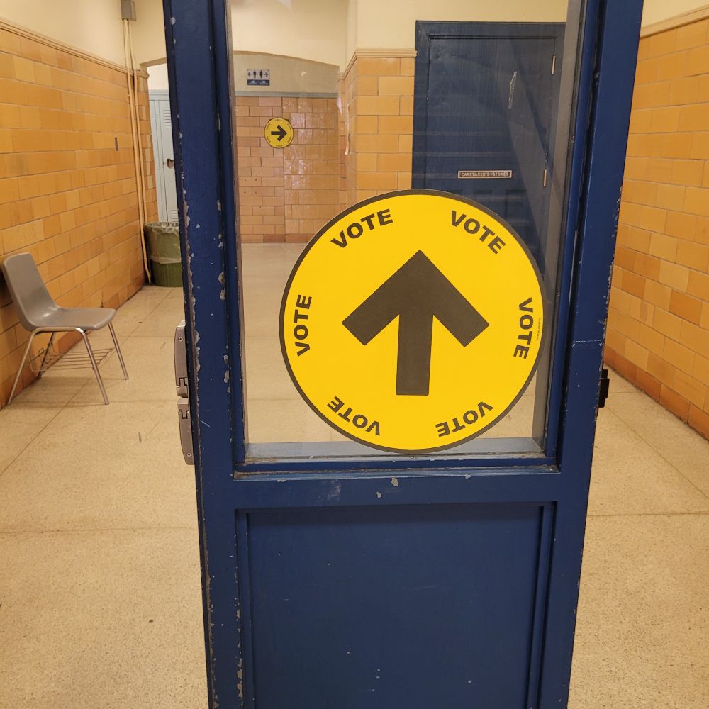 A yellow circular vote sign with a blush arrow in the middle. It is taped to a window in a blue doorway.