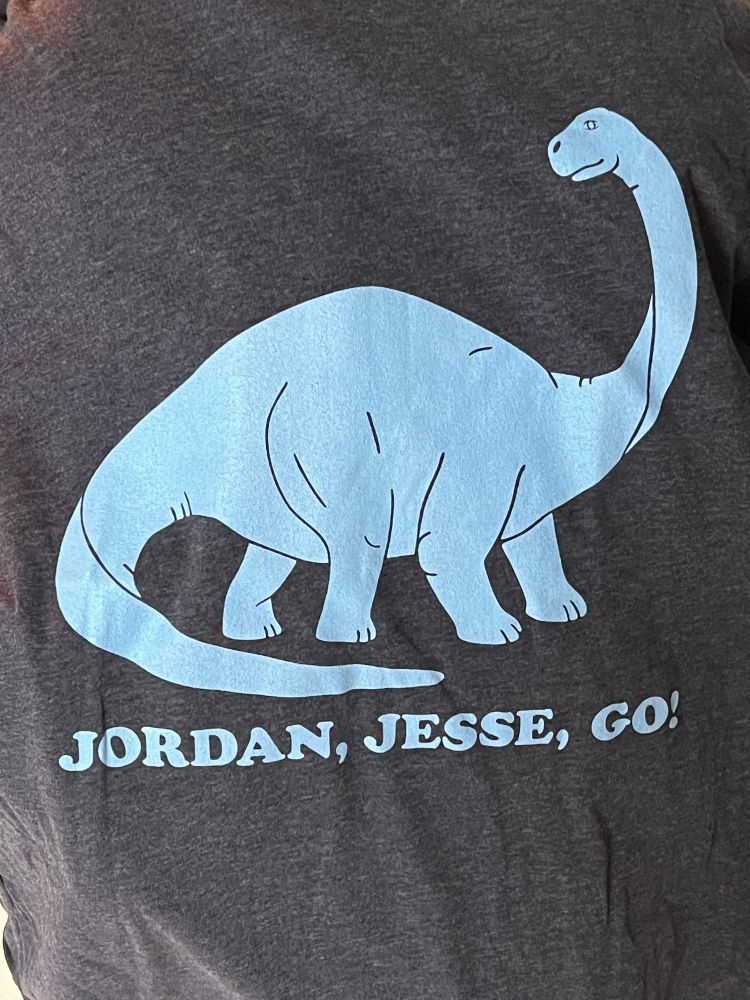 A dark Heather grey t shirt with a drawing of a brontosaurus above the text “Jordan, Jesse, go!” The dinosaur and text are light blue 