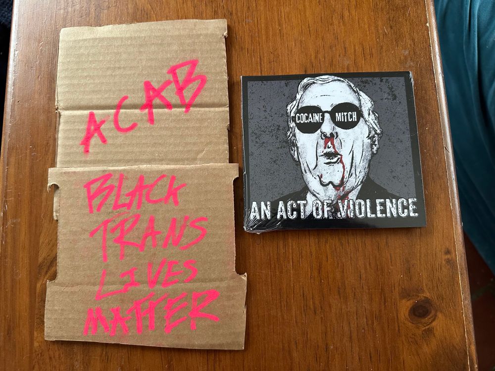 Photo of the cover of CD album “An Act of Violence” by Cocaine Mitch, showing Mitch McConnell in shades with a bloody nose. Next to it are two cardboard cushions that shipped with it. On the cardboard is written in hot pink marker “ACAB” and “Black trans lives matter.”