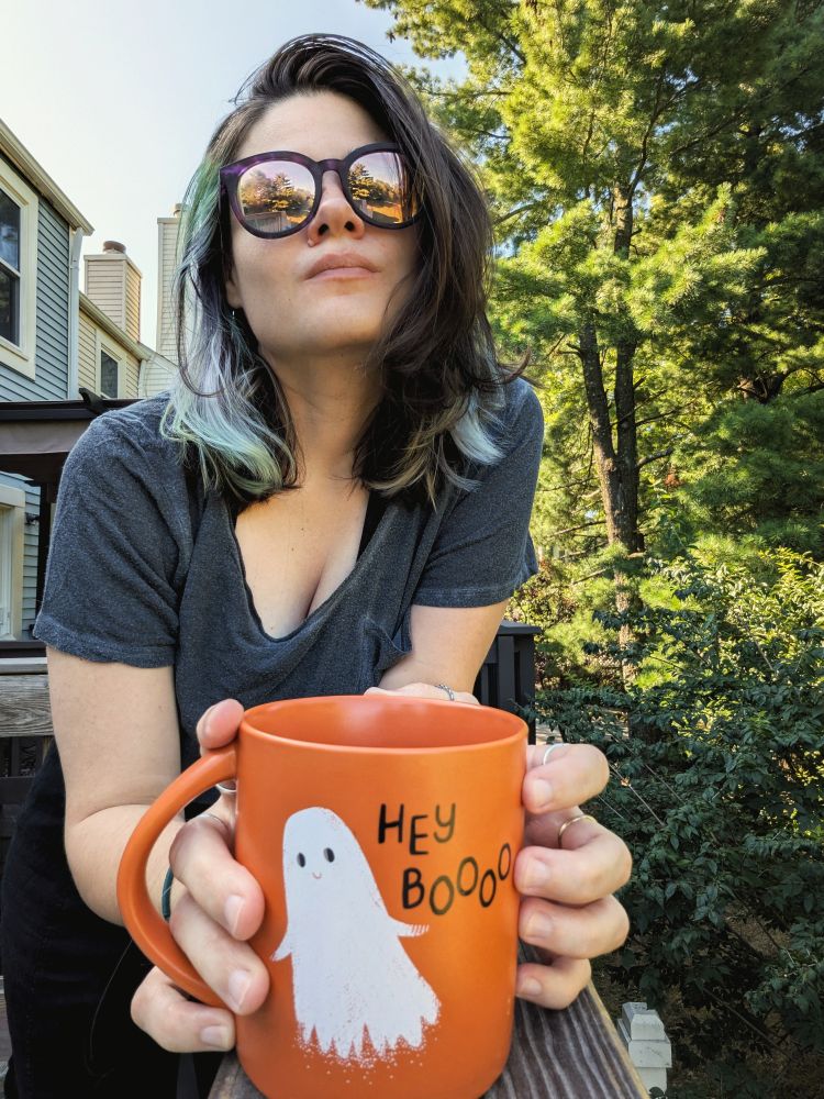 Andrea standing outside wearing sunglasses and holding an orange mug with a ghost on it that says "hey booo"