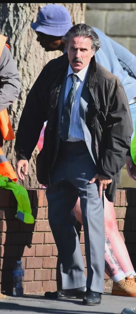 Con O'Neill in grey pants, a white shirt, a blue tie, and a black coat over a grey suit jacket.