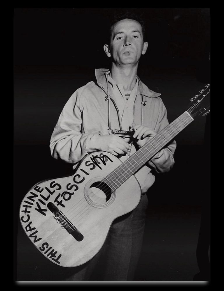 A photo of Woody Guthrie holding a guitar that says ‘This machine kills fascists’. Updated with very bad editing crossing out the letters ‘ts’ to now read ‘This machine kills fascism’.