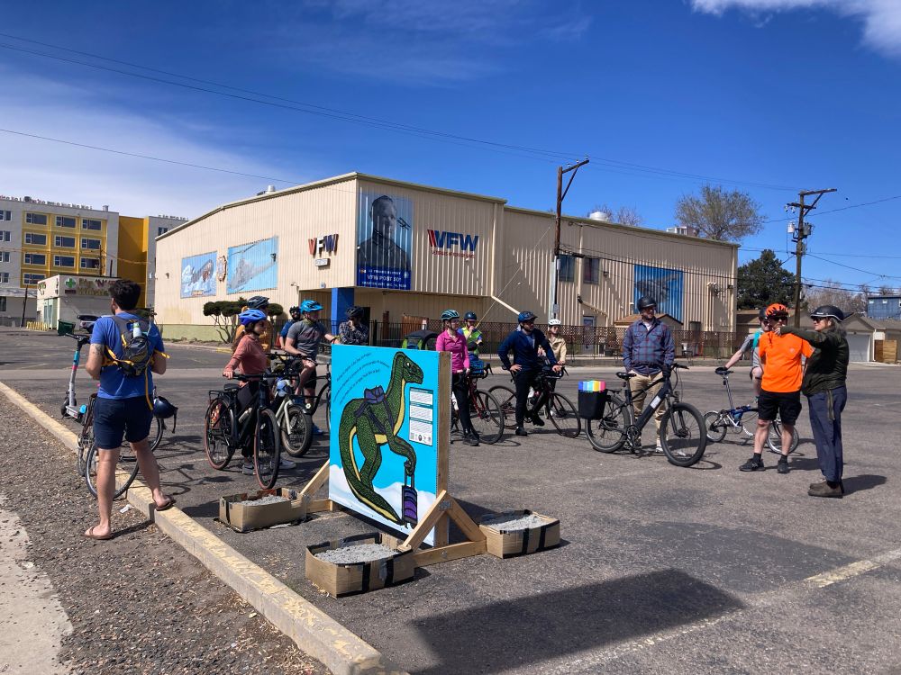 Photo of bike riders by a dinosaur painted on wood panel installed on a parking lot by bus stop with only a bench 