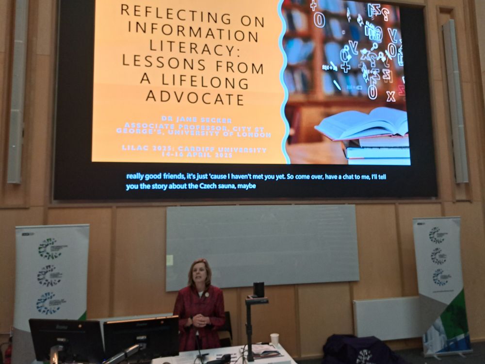 Jane Secker stood behind a podium at the front of the Sir Stanley Thomas lecture theatre in the Centre for Student Life (CSL) at Cardiff University. 

Behind Jane is a projection screen displaying a slide. On a dark yellow background, the slide says Reflecting on Information Literacy: Lessons from a Lifelong Advocate

Dr Jane Secker, Associate Professor, City St George's, University of London

LILAC 2025 University of Cardiff, 14-16 April

To the right of the text, there is a pile of books with an open book at the top and some white letters and characters above it. 
