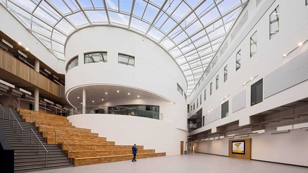 Inside The Wave at the University of Sheffield. The Wave is the Social Sciences building at the university. There is a large white circular structure with windows in the centre, to the right there is a white wall with windows, to the left there is tiered wooden seating and a grey staircase. The ceiling is glass and you can see the blue cloudy sky. 