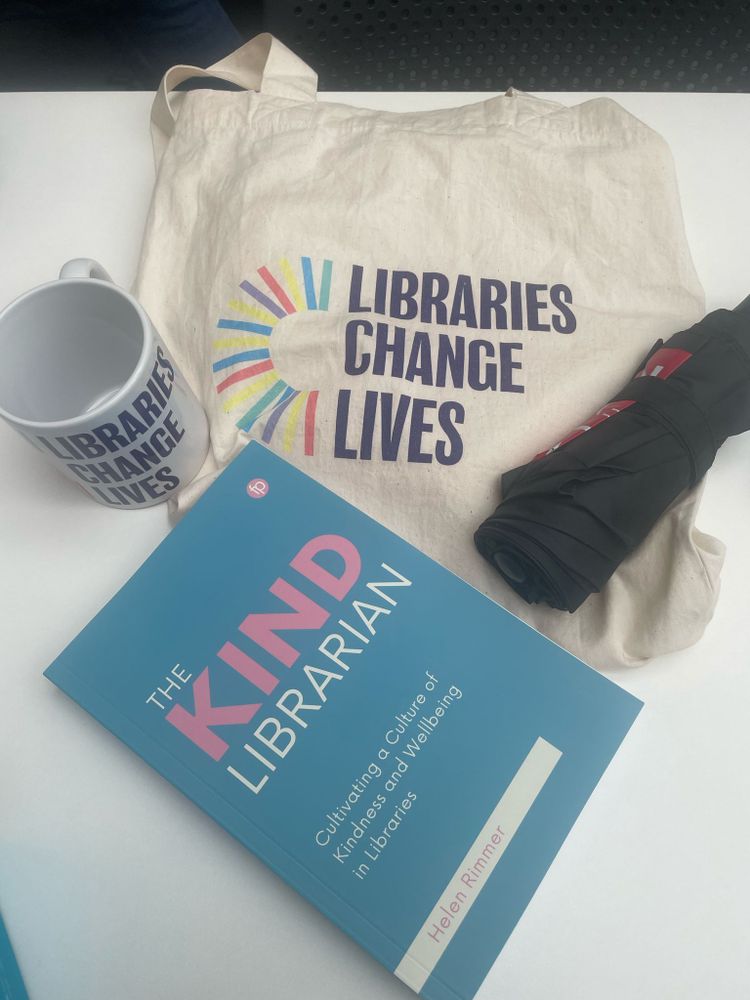 A combination of items on a white table. At the front is a book with the title 'The Kind Librarian', on top of a cream tote bag that says 'Libraries Change Lives'. To the right is a black umbrella and to the left is a white mug that says 'Libraries Change Lives'.