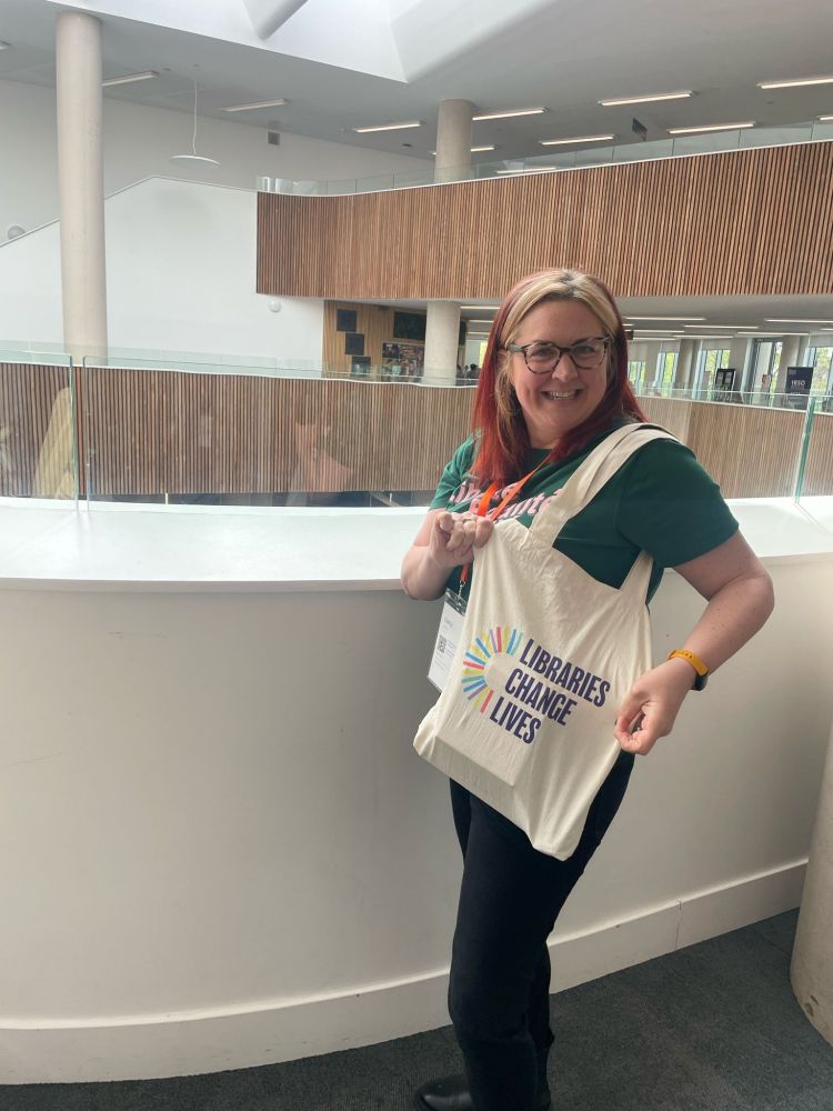 Lucy Collins holding a cream tote bag that says LIBRARIES CHANGE LIVES in uppercase letters that she won at the LILAC 2025 exhibition. 