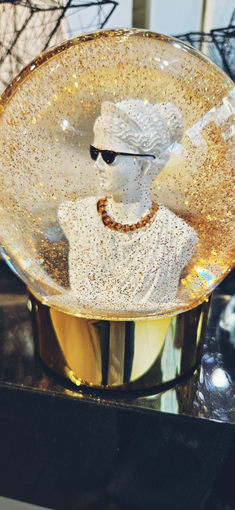 The image shows a glass snow globe filled with glittery, golden liquid and a female Grecian bust wearing sunglasses and a gold chain. The opaque globe is set against a dark background, creating a striking visual contrast.