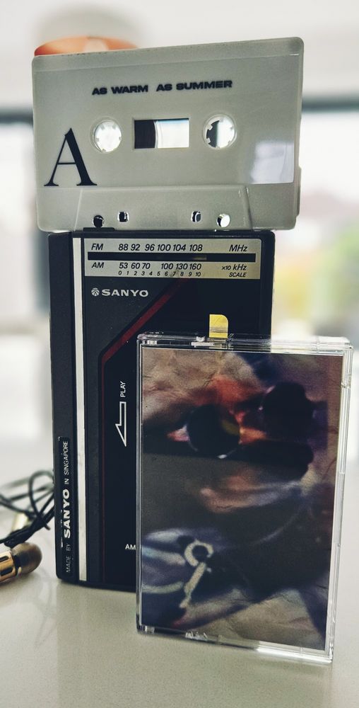 The image shows a nostalgic collection of vintage audio equipment. At the centre is a black Sanyo portable cassette player/radio (commonly known as a Walkman-style device) with visible FM/AM frequency scales (88-108 MHz for FM and 53-160 kHz for AM). 

There is a cassette tape in the image:
- A light gray cassette tape positioned on top with the text "AS WARM AS SUMMER" printed on it and the letter "A" indicating side A of the tape
- A clear plastic cassette case at the bottom containing what appears to be a cassette box, though the artwork on it is somewhat blurry, we can make out a dog wearing sunglasses.

In the bottom left corner of the image, you can see what looks like the plug end of headphones or earbuds with a gold-coloured connector.

This arrangement captures a slice of pre-digital music technology from the 1980s/1990s era, when portable cassette players and mix tapes were popular ways to listen to and share music.