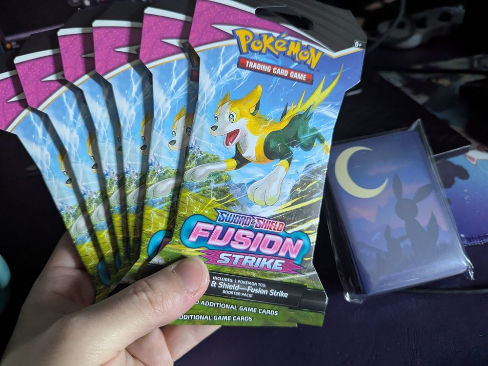 six Fusion Strike blister packs fanned in hand, with a pack of Umbreon & Eevee card sleeves in the background