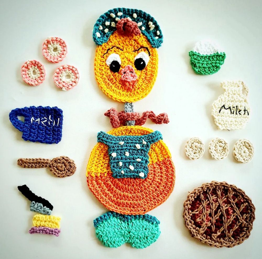 A remake of a vintage yellow duck in crochet. It’s a flatlay of the duck and  there are kitchen utilities also in crochet. A blueberry pie with a raster. Milkjug, cookies butter with a butterknife a wooden spoon a flowerpot and 3 egs
