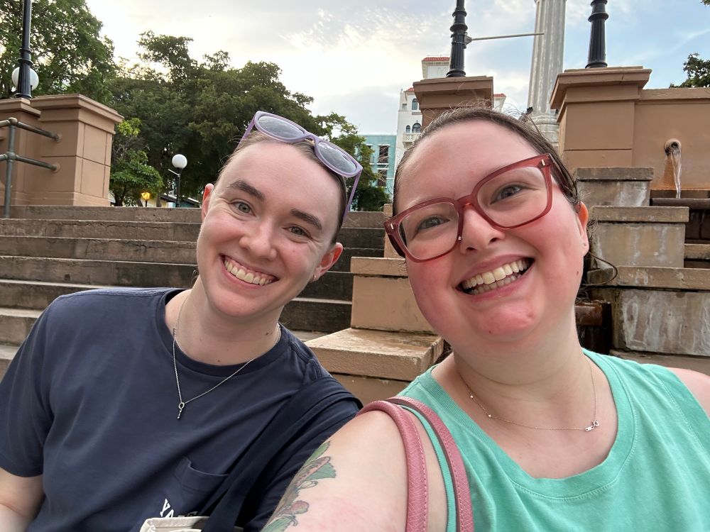 A selfie of Isabelle and Cassie, two young adult women, sitting in front of steps in Old San Juan. Both are smiling at the camera. 