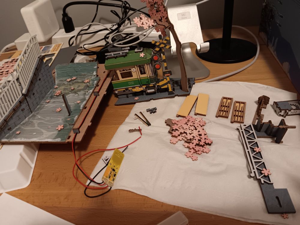 Some assembled parts laying next to more completed scenes. The bottom of the insert with the river sprinkled with cherry blossom petals and a railed staircase along side the river, the train on a track with the train signal and a cherry blossom tree, as well as individual parts of a few traditional Japanese shuttered windows, an AC unit, a cherry blossom branches, and a truss bridge railing. 