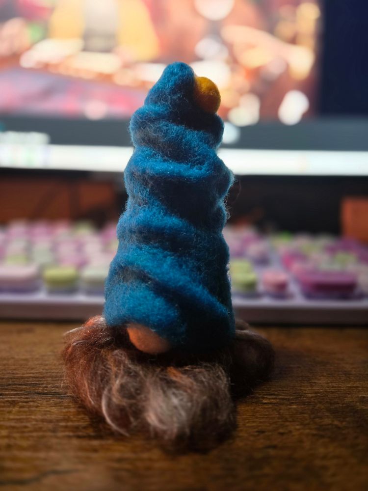 A needle felted gnome with a blue hat, in front of a key board and monitor.