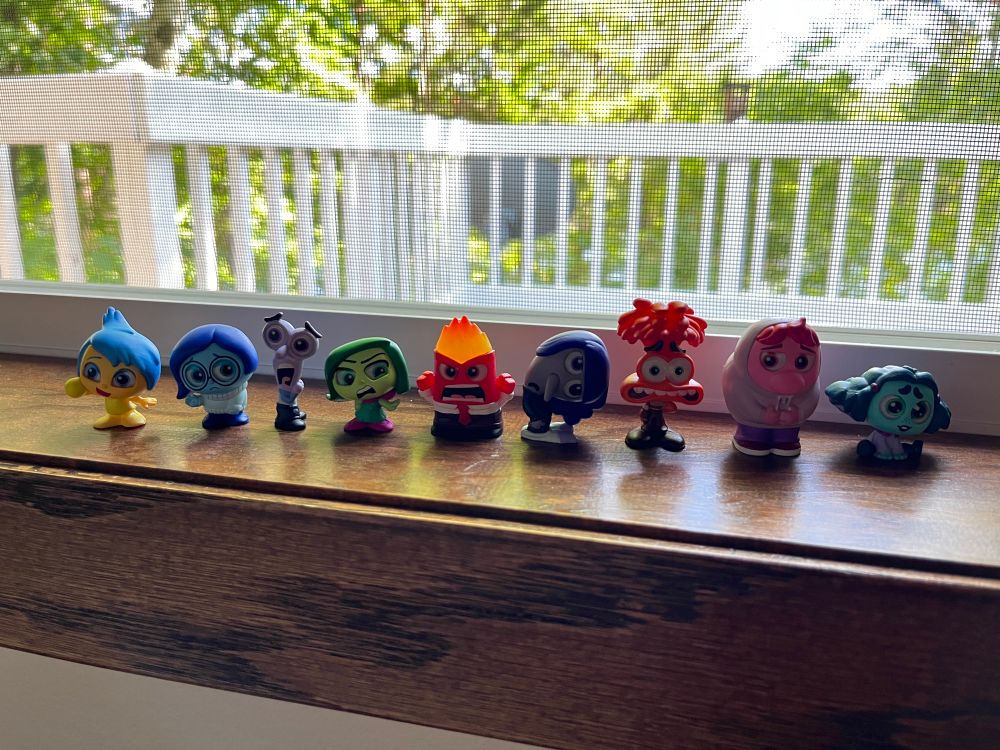 Disney doorable figures of the inside out 2 emotions; Joy, Sadness, Fear, Disgust, Anger, Ennui, Anxiety, Embarrassment and Envy sitting on a window sill. 