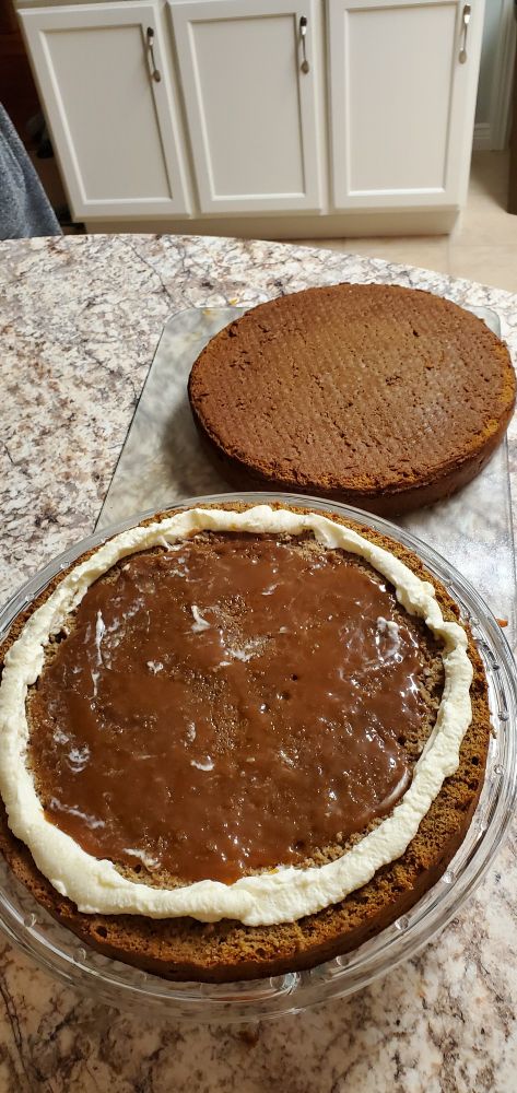 Two halves of a cake, one on a cake tray, the other beside it on the counter. Bottom half that's on the tray has a ring of icing around its outer edges and has caramel within that ring. The top half next to this was soaked with liquid and as such looks wet.