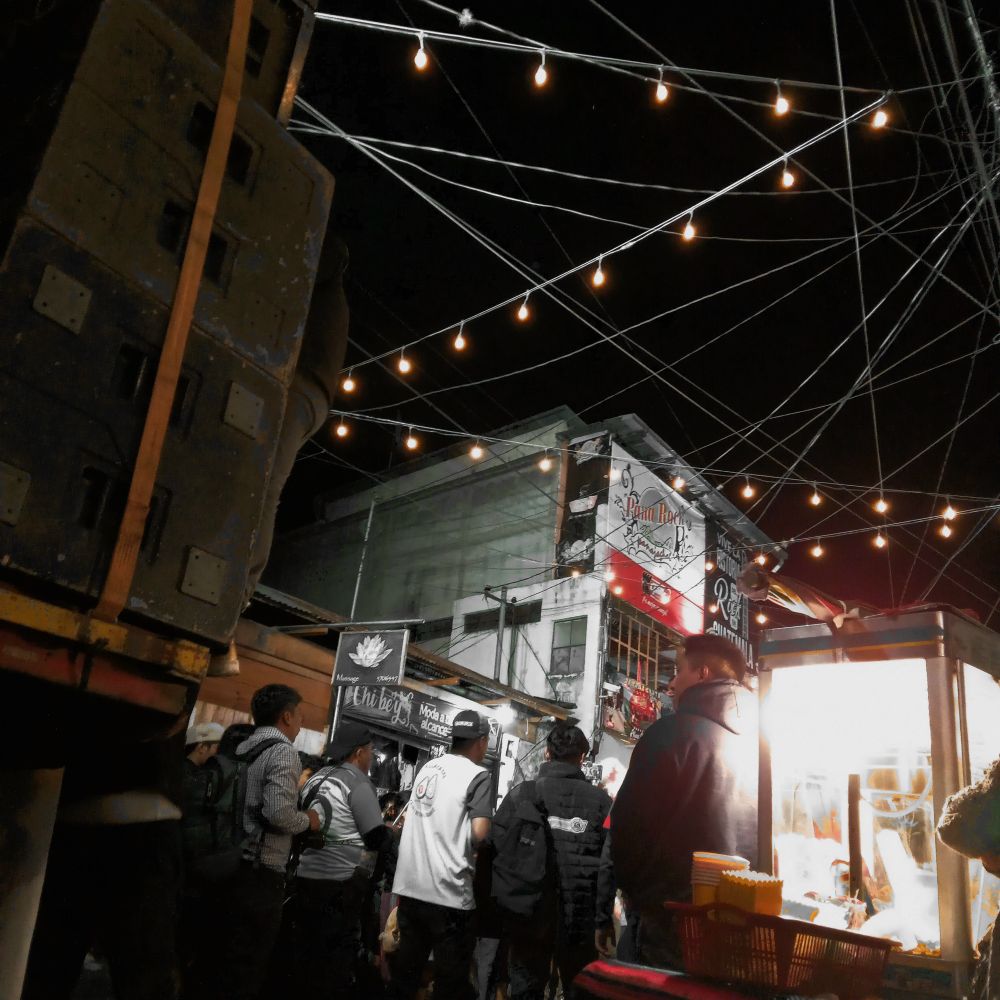 Popcorn vendor and group of men next to the El Chavo themed semi-truck carrying a rave calliber speaker stack. Hanging lights and a nest of power lines above the street.