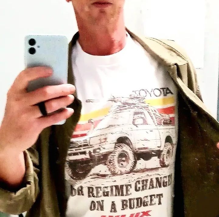 Guy taking a selfie wearing a white printed t-shirt with an illustration of a lifted Toyota Hilux 4x4 with a missile launcher mounted in the bed. Text reads: Toyota Hilux: For Regime On a Budget"