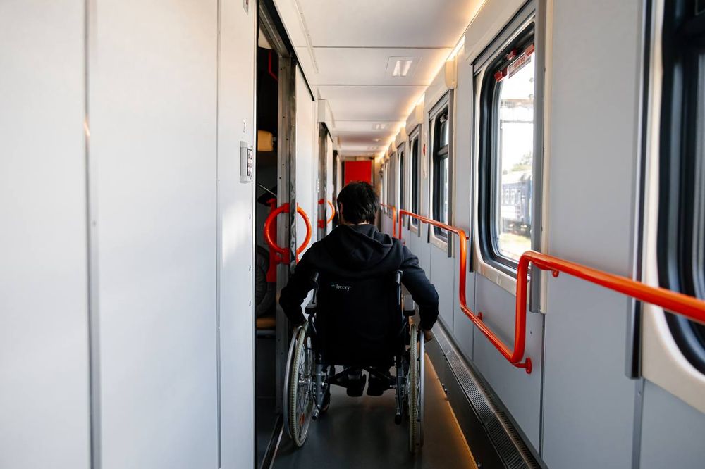 a view from behind of a person in dark clothing rolling a wheelchair down the corridor of a rail car with white walls and bright orange railings and door handles