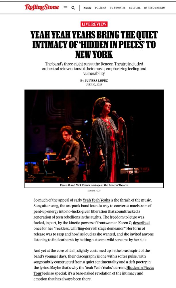 Screenshot of the Rolling Stone review. Rolling Stone logo appears at the top menu followed by website options. Underneath that is a red box w/ white text states LIVE REVIEW. Underneath the Live Review box is the title of the review, "Yeah Yeah Yeahs Bring the Quiet Intimacy of 'Hidden in Pieces' to New York. More text under that reads: The band’s three-night run at the Beacon Theatre included orchestral reinventions of their music, emphasizing feeling and vulnerability.
Article is by Julyssa Lopez published July 30, 2025. 

Underneath this is an image by me (Edwina Hay) of Nick Zinner in the background playing acoustic guitar w/ a music stand in front of him. Karen O is in the foreground wearing a headpiece, that is gold and resembles a snake. She is singing wearing a blue beaded cape over a red jumpsuit. Underneath the image is a caption that states: Karen O. and Nick Zinner onstage at the Beacon Theatre. 

The first two paragraphs of the review follow: So much of the appeal of early Yeah Yeah Yeahs is the thrash of the music. Song after song, the art-punk band found a way to convert a maelstrom of pent-up energy into no-fucks-given liberation that soundtracked a generation of teen rebellions in the aughts. The freedom to let go was fueled, in part, by the kinetic powers of frontwoman Karen O, described once for her “reckless, whirling-dervish stage demeanor.” Her form of release was to rasp and howl as loud as she wanted, and she invited anyone listening to find catharsis by belting out some wild screams by her side.  

And yet at the core of it all, slightly costumed up in the brash spirit of the band’s younger days, their discography is one with a softer pulse, with songs subtly constructed from a quiet sentimentality and a deft poetry in the lyrics. Maybe that’s why the Yeah Yeah Yeahs’ current Hidden in Pieces Tour feels so special; it’s a bare-naked revelation of the intimacy and emotion that has always been there.



